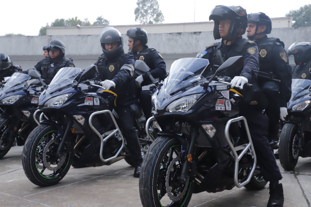Eine Gruppe von Polizisten in schwarzen Uniformen, Helmen und Handschuhen, die Motorräder in einer Parkanlage mit Bäumen, einer Wand und einem klaren blauen Himmel im Hintergrund fahren.