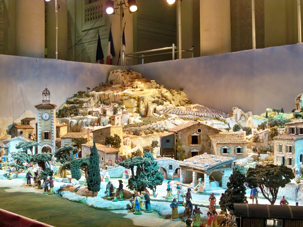Eine Museumsschau einer Krippe mit Menschen, Häusern, Bäumen, Felsen, einem Kirchturm, Fahnen, Pfählen und Lichtern, alles beleuchtet und eine festliche Atmosphäre schaffend.