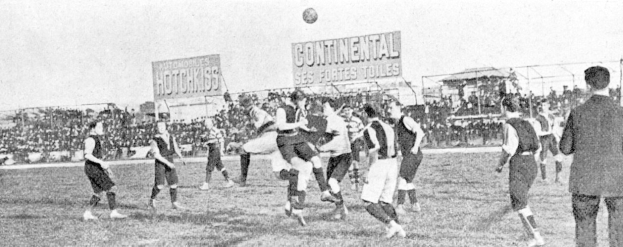 Ein Schwarz-Weiß-Bild einer Gruppe von Männern, die auf einem Feld Fußball spielen, mit einem Ball in der Luft und Zuschauern im Hintergrund, mit Tafeln, auf denen Informationen zum Spiel angezeigt werden.