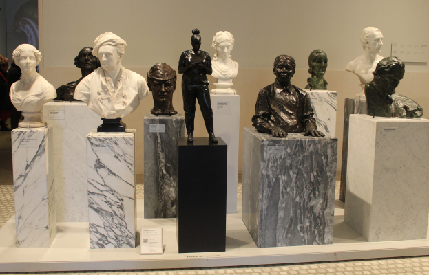 Eine Gruppe detaillierter, lebensechter Büsten-Skulpturen im Museum ausgestellt, mit ein paar Menschen im Hintergrund und einer Wand dahinter.