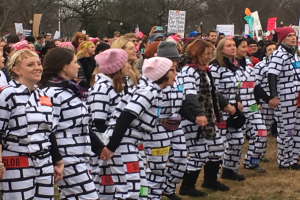 Eine Gruppe von Frauen in Gef√§ngnisuniformen steht in einer Reihe auf einer grasbewachsenen Fl√§che, h√§lt Schilder und tr√§gt Kappen, mit B√§umen und einem klaren blauen Himmel im Hintergrund.