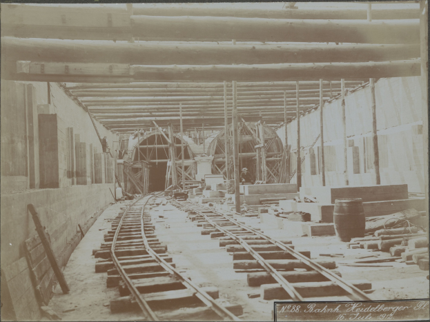 Schwarz-weiß-Foto einer Eisenbahnschiene in einem Gebäude unter Bau, mit verstreuten Holzgegenständen, Fässern, Wänden, Pfählen und Text unten.