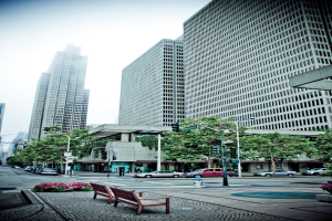 Eine Außenansicht mit einer Straße, auf der Autos geparkt sind, flankiert von Bänken mit kleinen Pflanzen, Bäumen und Hochhäusern im Hintergrund.