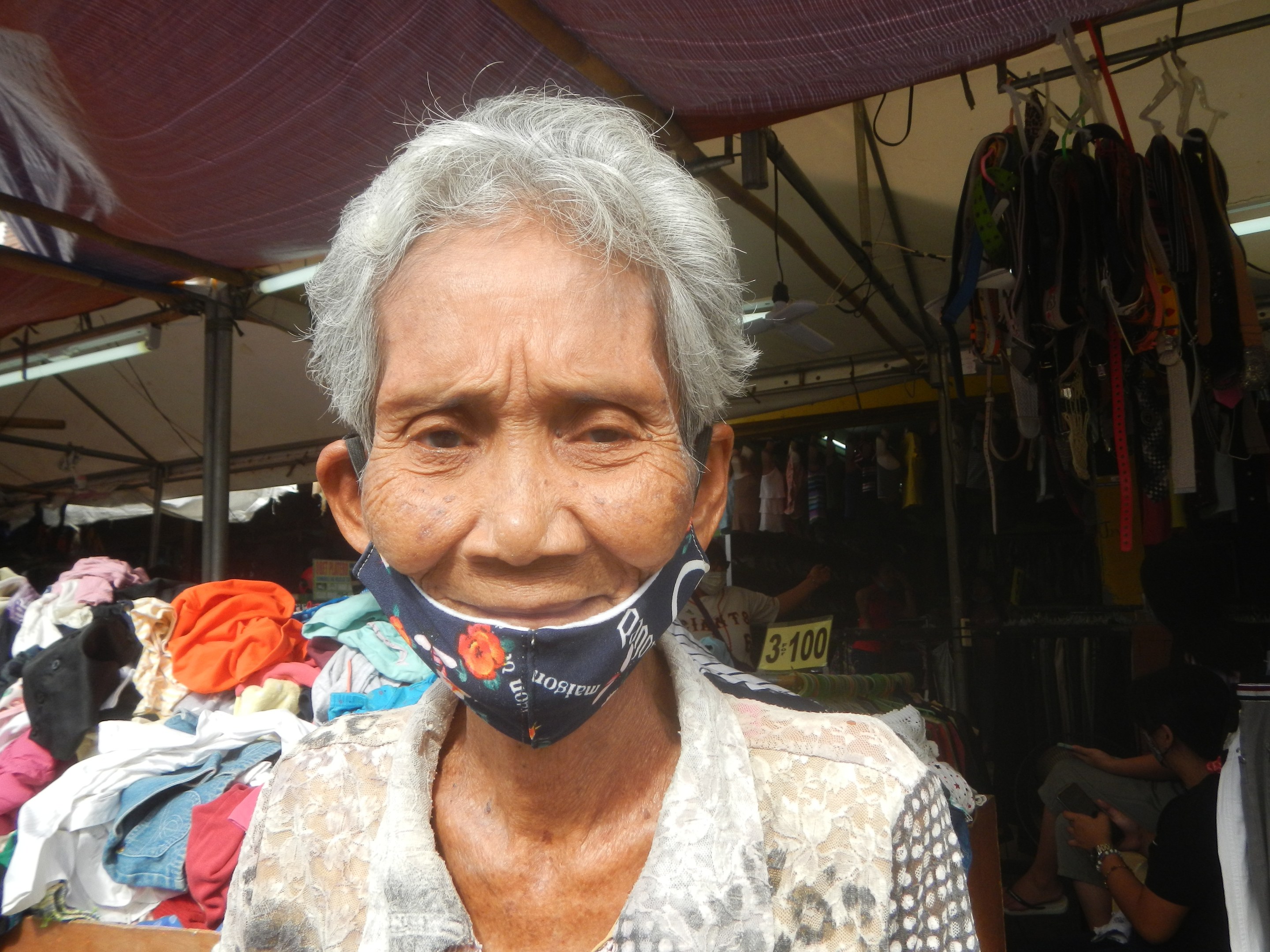 Eine ölte Frau mit Gesichtsmaske steht auf einem Markt umgeben von hängenden Klamotten, Lichtern, Ventilatoren und anderen Gegenständen, mit einer Person im Hintergrund sitzend.