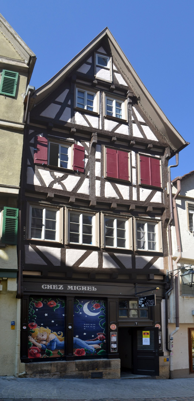 Altes Gebäude mit roten Fensterläden, mehreren Fenstern und klassischem Design, das sich auf einer Straße mit einem klaren blauen Himmel im Hintergrund befindet.
