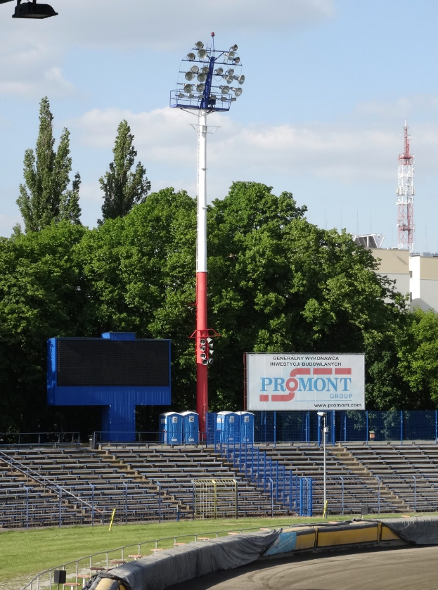 Eine von Scheinwerfern beleuchtete Rennbahn, umgeben von Gras, Geländern, Stufen, einem Lichtmast, einem Schild, Bäumen, Gebäuden, Türmen und einem bewölkten Himmel.