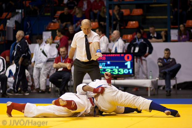 Zwei Judokämpfer im Kampf, mit einem Schiedsrichter in der Nähe, in einer Halle mit Sitz- und Stehplätzen, Geländern, Treppen und einer Anzeigetafel im Hintergrund.