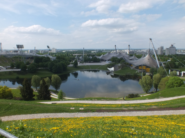 Blick auf das Olympiastadion von einem Hügel aus, mit einem Teich im Vordergrund, der von Grünflächen und gelben Blumen umgeben ist, und Gebäuden im Hintergrund unter einem klaren blauen Himmel.