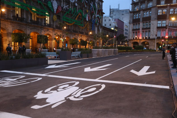 Stadtstraße mit einem ausgewiesenen Fahrradweg, umgeben von Gebäuden, Straßenmöbeln, Fußgängern auf dem Gehweg und Grün unter einem klaren blauen Himmel.