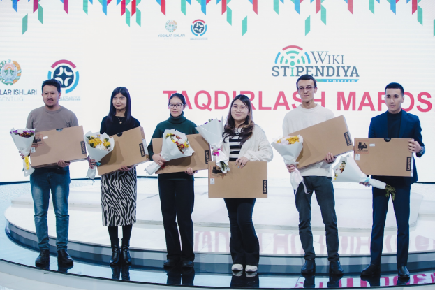 Gruppe von Menschen auf einer Bühne mit Blumensträußen und Kartons, im Hintergrund ein Banner mit der Aufschrift "Yoslar Ishlari" und "Wikki Stipendiya"