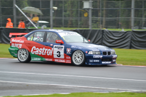 Ein Auto fährt auf einer Rennstrecke mit Text und Zahlen darauf, umgeben von Gras, mit Menschen in orangen Jacken, die Schirme halten und Bäumen im Hintergrund hinter einem Maschendrahtzaun, der mit schwarzem Stoff bedeckt ist.