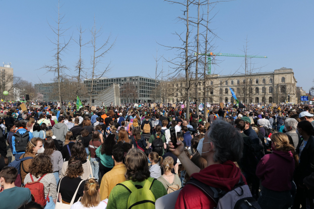 Eine große Menge von Menschen mit Schildern und Tötenschutzwesten, die vor einem Gebäude mit Bäumen und einem klaren Himmel im Hintergrund stehen, was auf eine Klimawandel-Demonstration hindeutet.