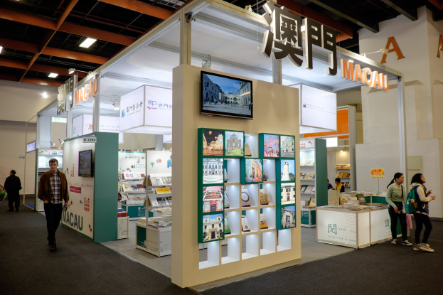 Menschen vor einem Messestand auf der Macau International Book Fair, gefüllt mit Büchern, einem montierten Fernseher, Namensschildern und beleuchteten Displays.