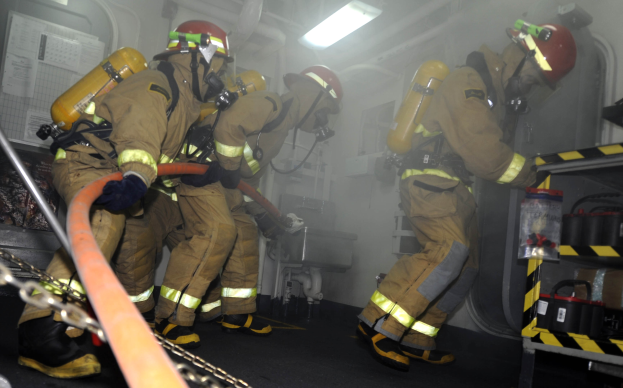 Feuerwehrleute in Schutzausrüstung mit Helmen und Sauerstoffflaschen arbeiten an einem Feuerwehrauto, mit einem Gestell voller Gegenstände und Hintergrundausrüstung im Hintergrund.