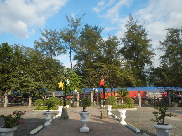 Ein Park mit Topfpflanzen und Bäumen in der Mitte, umgeben von Straßenlaternen, Dekorationen und Grills unter einem bewölkten Himmel.