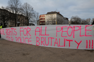 Eine Gruppe von Menschen, die draußen vor einem Banner mit der Aufschrift 'Rechte für alle Menschen Stoppt Polizeigewalt' stehen, mit Straßenlaternen, einem Schild, Bäumen, Gebäuden und einem bewölkten Himmel im Hintergrund.