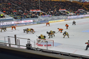 Gruppe von Menschen, die Hockey auf einem Eisring mit Netz und Umzäunung spielen, Zuschauern im Hintergrund, Bannern mit Text, Stadionbeleuchtung