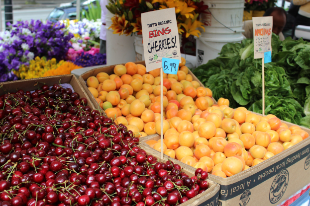 Ein Bauernmarkt mit Kisten voller Kirschen, Orangen und Blattgemüse, Schilder mit Text und Menschen, Fahrzeugen, Pfählen und Gebäuden im Hintergrund.