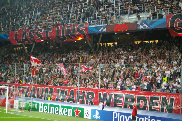 Ein Fußballspiel in einem Stadion mit einer großen Zuschauermenge, saftig grünem Rasen, einem Torpfosten links, Bodenbannern, Menschen mit Fahnen und Hintergrundgeländern, Tafeln, Lichter und andere Gegenstände.