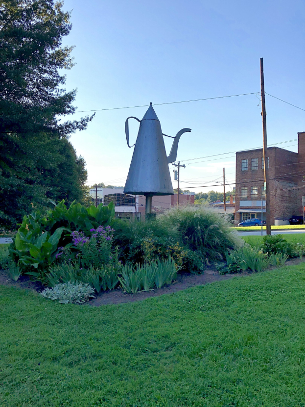 Ein Metallgießer steht auf einem grünen Feld mit Pflanzen, Bäumen, Strommasten, einem Schild, Fahrzeugen, Gebäuden und einem bewölkten Himmel im Hintergrund.