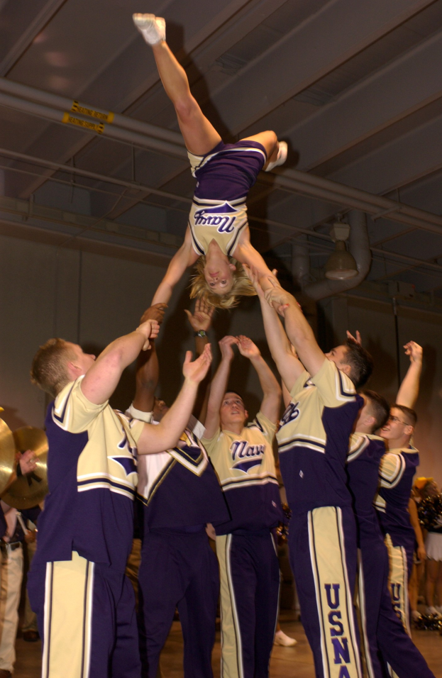 Eine Gruppe von Cheerleadern führt einen Stunt in einer Turnhalle durch, wobei eine Frau in einem Handstand in der Mitte steht und von anderen umgeben ist, die Musikinstrumente halten, während Rohre und Deckenlampen im Hintergrund zu sehen sind.