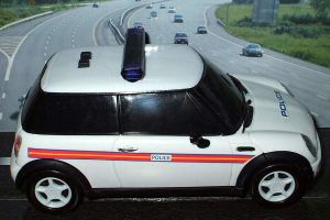 Ein weißes Polizeifahrzeug ist im Vordergrund geparkt, hinter dem sich mehrere Fahrzeuge auf der Straße bewegen, flankiert von Gras und Bäumen auf beiden Seiten in einer ländlichen Umgebung.