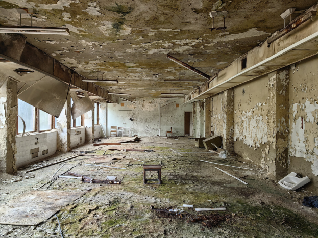 Verlassener Krankenhausinnenraum mit abblätternder Farbe, verstreuten Trümmern einschließlich Holz und einem Hocker sowie Fenstern, die natürliches Licht auf der linken Seite einlassen.